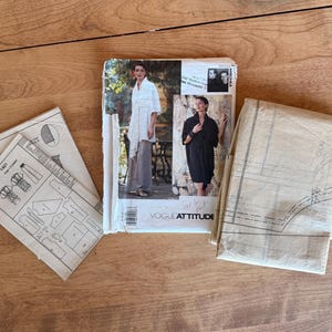 May include: A collection of vintage Vogue sewing patterns, including a pattern booklet and several pattern sheets. The main pattern features a woman in a white top and gray pants. The patterns are on a wooden surface.