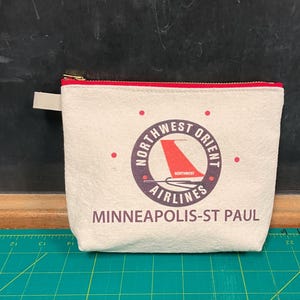 May include: A white canvas zippered pouch with a red zipper pull. The pouch has a vintage logo for Northwest Orient Airlines with a red airplane wing and the text "Northwest Orient Airlines" in a circle. Below the logo is the text "Minneapolis-St. Paul".