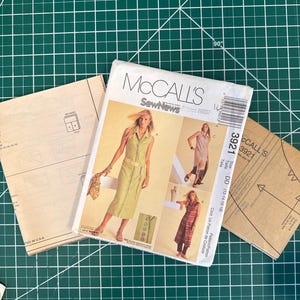 May include: A McCall's sewing pattern packet with images of a woman wearing a dress and a long coat. The packet is on a green cutting mat, with two pattern sheets visible. The packet has the text "McCall's SewNews".