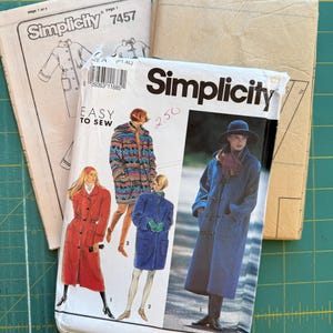 May include: A collection of vintage Simplicity sewing patterns. The main pattern features illustrations of coats in red, blue, and a patterned design. The text on the pattern reads "Simplicity 7457" and "Easy to Sew". The background is a green cutting mat.