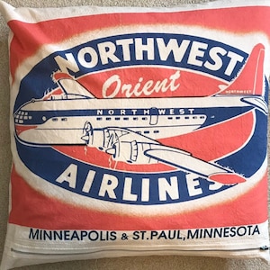 May include: A vintage-style pillow featuring a red, white, and blue logo for Northwest Orient Airlines. The logo depicts a plane flying with the words "Northwest Orient Airlines" and "Minneapolis & St. Paul, Minnesota" in white text on a red background.