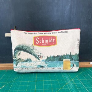 May include: A white canvas zippered pouch with a vintage Schmidt Beer advertisement. The ad features a leaping fish, a glass of beer, and the text "The Brew that Grew with the Great Northwest" and "Schmidt Beer".