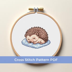 May include: A cross-stitch pattern of a sleeping hedgehog on a blue pillow, framed in a wooden embroidery hoop. The hedgehog is brown and tan, with closed eyes. The text "Cross Stitch Pattern PDF" is displayed on a blue banner.