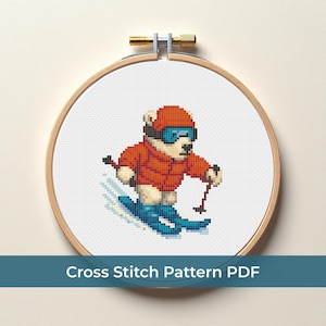 May include: Cross-stitch design of a polar bear skiing, wearing an orange jacket, blue goggles, and a matching helmet. The bear is on blue skis with black poles. The design is in a wooden embroidery hoop. Text at the bottom reads "Cross Stitch Pattern PDF".