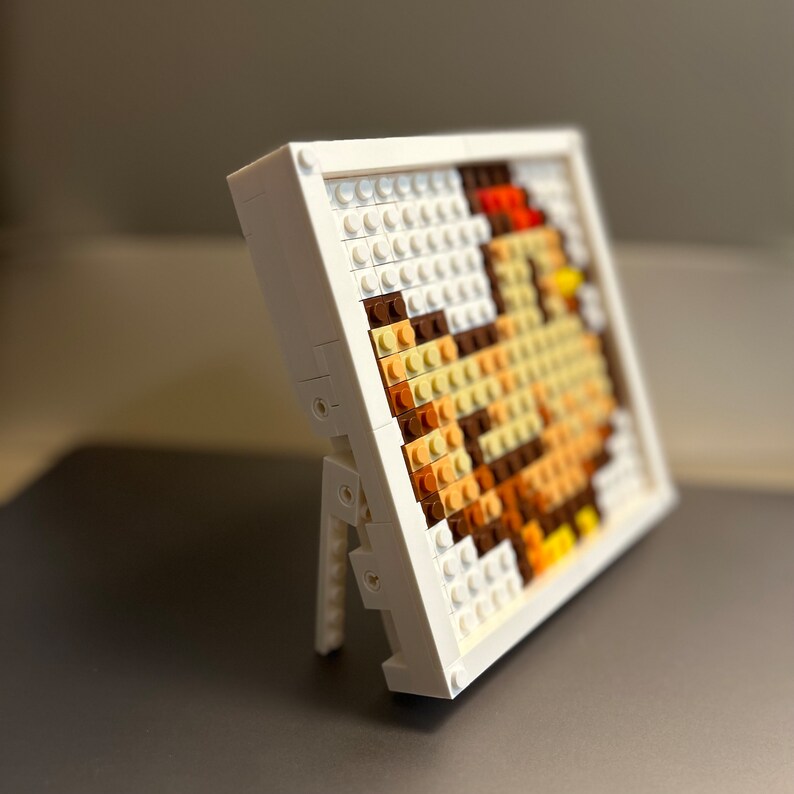 Op de afbeelding: Een wit frame met een standaard houdt een moza&iuml;ekbeeld van een haan vast, gemaakt van kleine plastic blokken in tinten bruin, oranje en wit.