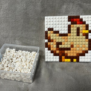 Op de afbeelding: Een witte plastic container gevuld met kleine witte plastic blokken. Naast de container is een vierkante moza&iuml;ek van een bruine kip gemaakt van kleine plastic blokken.