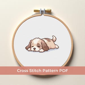 Könnte beinhalten: Ein Kreuzstichmuster eines schlafenden Welpen in einem hölzernen Stickrahmen. Der Welpe ist hellbraun und weiß, mit dunkelbraunen Akzenten. Der Text "Cross Stitch Pattern PDF" wird darunter angezeigt.
