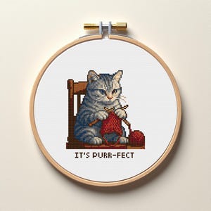 May include: Cross-stitch embroidery in a wooden hoop features a gray tabby cat knitting with red yarn. The cat sits on a wooden chair, with the text "IT'S PURR-FECT" below the cat.