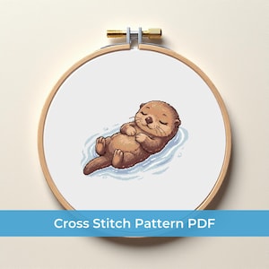 Peut inclure: Un motif de point de croix représentant une loutre de dessin animé flottant sur l'eau. La loutre est brune, les yeux fermés et l'expression paisible. Le motif est encadré dans un cercle à broder en bois. Le texte en bas indique "Cross Stitch Pattern PDF".