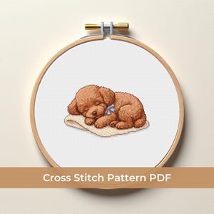 May include: A cross-stitch pattern of a sleeping brown poodle puppy with a blue bow, resting on a beige blanket. The design is displayed in a wooden embroidery hoop. Text at the bottom reads "Cross Stitch Pattern PDF".