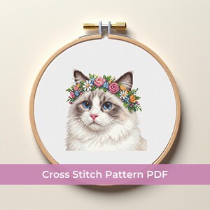 Puede incluir: Un patrón de punto de cruz en PDF que muestra un gato blanco y gris con ojos azules que lleva una corona floral colorida de flores rosas, amarillas y azules, exhibido en un bastidor de madera redondo sobre tela blanca.