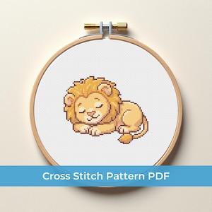 May include: A cross-stitch pattern of a sleeping lion in a wooden embroidery hoop. The lion is light brown with a yellow mane and closed eyes. The text "Cross Stitch Pattern PDF" is in a blue banner at the bottom.