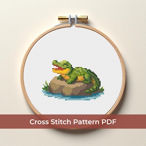 Könnte beinhalten: Kreuzstichmuster eines pixeligen grünen Alligators, der auf einem braunen Felsen im Wasser liegt. Das Design ist in einem hölzernen Stickrahmen eingefasst. Der Text lautet "Cross Stitch Pattern PDF".