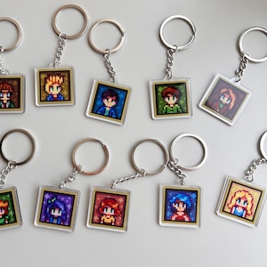 May include: A collection of keychains featuring pixelated character portraits. Each keychain has a silver ring and chain, with a square, clear acrylic pendant. The portraits showcase various characters with different hair colors and outfits, set against a gold border.