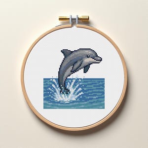 Op de afbeelding: Een borduurring met kruissteek met een gepixeleerde dolfijn die uit blauw water springt. De dolfijn is grijs en wit, met een gedetailleerd ontwerp. De ring is gemaakt van hout en heeft een messingkleurig aanspanningsmechanisme.