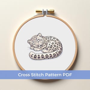 May include: A cross-stitch pattern of a sleeping snow leopard, featuring a white body with black spots, resting on a white background. The design is framed in a wooden embroidery hoop. Text at the bottom reads "Cross Stitch Pattern PDF".