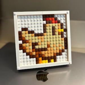 Op de afbeelding: Een wit frame met een moza&iuml;ek van kleurrijke plastic vierkanten die een kip voorstellen. De kip is bruin, geel en oranje.