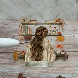 Op de afbeelding: Een aquarel-illustratie van een vrouw met lang bruin haar die aan een bureau zit met een boek en een kop koffie. De vrouw draagt een beige trui en een bloemenband in haar haar. De illustratie is omringd door herfstachtige elementen, zoals pompoenen, bladeren en bloemen.