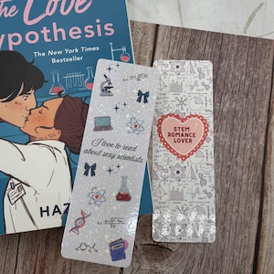 May include: A stack of items includes a paperback book titled "The Love Hypothesis" and two decorative bookmarks. One bookmark has the text "I love to read about sexy scientists" and the other says "STEM Romance Lover". The bookmarks have science-themed illustrations.