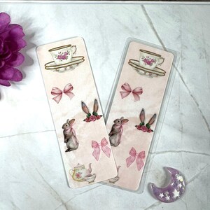 Peut inclure: Deux marque-pages roses et blancs avec une tasse et une soucoupe, une théière, un lapin et des nœuds roses. Les marque-pages sont sur une surface blanche avec une lune croissante violette.