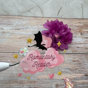 Peut inclure: Un autocollant transparent avec le texte "Romantasy Reader" en rose, représentant une silhouette de dragon noir, des nuages roses, des étoiles jaunes et des cœurs roses. Une fleur violette et un croissant de lune rose pailleté sont également présents.