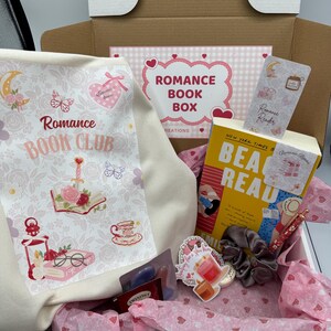 May include: A Romance Book Club box filled with a book, scrunchies, stickers, tea, and a pen. The box includes a canvas bag with the words "Romance Book Club" and a decorative design. The book has a yellow cover with the title "BEA READ".