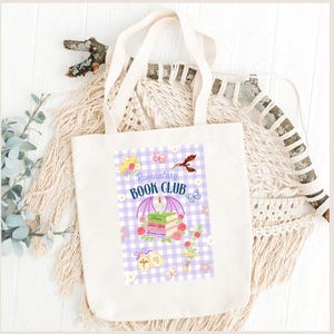 Peut inclure: Sac fourre-tout crème avec l'inscription "Romantasy Book Club" et un motif de livres, de fleurs et d'un dragon sur un fond à carreaux vichy violets. Le sac a deux anses et est fait d'un tissu résistant.
