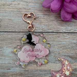 Peut inclure: Un porte-clés en acrylique transparent avec un fermoir en forme de cœur en or rose. Le porte-clés présente un motif de nuage rose avec les mots "Romantasy Reader" en cursive, une silhouette de chauve-souris noire et des accents d'étoiles et de cœurs.