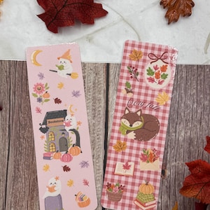Peut inclure: Deux marque-pages décoratifs avec des illustrations sur le thème de l'automne. L'un représente une librairie, des citrouilles et un fantôme. L'autre présente un renard, des feuilles et le texte "Cozy Fall" sur un fond à carreaux rouges et blancs.