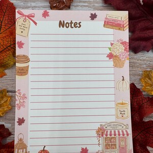 Peut inclure: Un bloc-notes rectangulaire avec des lignes horizontales et le mot Notes en haut. La bordure pr&eacute;sente des illustrations de livres, une tasse de caf&eacute; marqu&eacute;e Hello Autumn, une citrouille, une bougie marqu&eacute;e Vanilla Maple et une boutique Cozy Chapters.