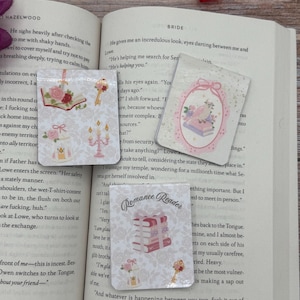 Peut inclure: Trois aimants rectangulaires décoratifs sur le thème romantique sont affichés sur un livre ouvert. Les aimants présentent des illustrations de livres, de roses, de clés et de bougies dans des tons rose et blanc doux. Un aimant lit "Romance Reader."