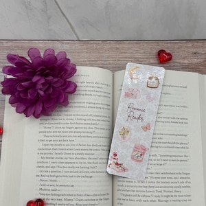 Peut inclure: Un marque-page décoratif avec le texte "Romance Reader" et des illustrations de fleurs, de papillons et d'un pot de confiture. Le marque-page est posé sur un livre ouvert avec une fleur violette et des décorations en forme de cœur.