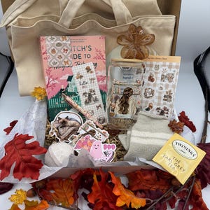 Peut inclure: Un coffret cadeau rempli d'articles sur le thème de l'automne, dont un livre intitulé "Witch's Guide to Magical Thinking", des autocollants, un bocal en verre, un sac fourre-tout beige et un sachet de thé. Le coffret est décoré de feuilles d'automne.