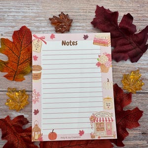 Peut inclure: Un bloc-notes rectangulaire avec des lignes horizontales et le mot Notes en haut. La bordure pr&eacute;sente des illustrations de livres, une tasse de caf&eacute;, une citrouille, une bougie, une lanterne et une boutique nomm&eacute;e Cozy Chapters. Le texte Holy & Joy Creations est en bas.