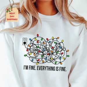 May include: White sweatshirt with a graphic of tangled Christmas lights plugged into an outlet. The text "I'M FINE. EVERYTHING IS FINE." is printed below the lights in black. The sweatshirt is a casual, comfortable style.