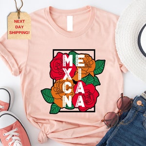 May include: A light pink t-shirt featuring the word "MEXICANA" in white block letters, framed by a black rectangle and colorful roses. The roses are red, orange, and yellow with green leaves. The shirt is paired with pink sneakers, blue jeans, and a white hat.