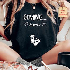 Op de afbeelding: Een zwart t-shirt met witte tekst die COMING... soon leest, samen met twee kleine hartjes. Onder de tekst staan twee babyvoetjes met hartvormige hielen, met de woorden OCTOBER en 2024 onder de voetafdrukken gedrukt.
