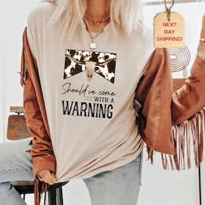 May include: A beige t-shirt with a cow skull graphic and the text "Should've come with a warning". The model is wearing the shirt with a brown suede jacket with fringe.