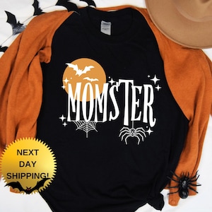 May include: Black and orange long-sleeved shirt with a white graphic of a moon, bats, a spider web, and the text "MOMSTER".