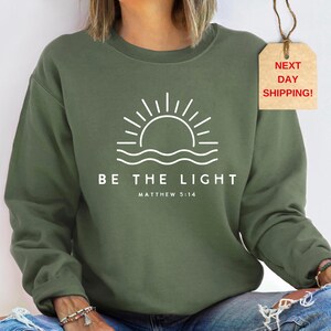 May include: A green sweatshirt with a white graphic of a sunrise over water and the text "BE THE LIGHT" with the scripture reference "MATTHEW 5:14".