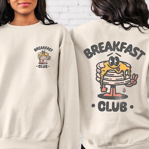May include: A cream-colored sweatshirt with a cartoon pancake graphic and the text "Breakfast Club" on the back. The front of the sweatshirt has a smaller version of the graphic and text.
