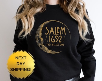 May include: Black sweatshirt with a gold graphic of a crescent moon and the words "SALEM 1692" and "THEY MISSED ONE". The sweatshirt has a round neck and long sleeves. The image also includes a gold starburst sticker that says "NEXT DAY SHIPPING!"