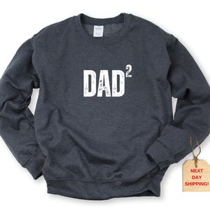 May include: Dark gray sweatshirt with the word "DAD²" printed in white across the chest. The sweatshirt has a crew neck and long sleeves. A tag with the text "NEXT DAY SHIPPING!" is attached to the garment.