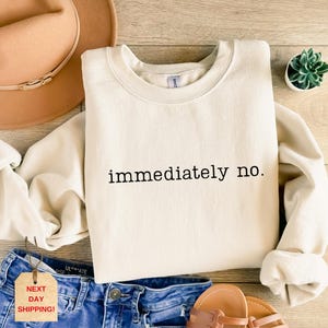 May include: Cream-colored sweatshirt with the phrase "immediately no" in black lettering. The sweatshirt is laid flat on a wooden surface, next to a pair of blue jeans, a brown hat, and sandals. A small plant in a white pot is also visible.