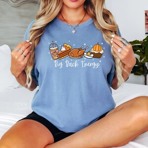 Puede incluir: Camiseta azul claro con un gr&aacute;fico de alimentos de Acci&oacute;n de Gracias, que incluye un caf&eacute; con leche de calabaza especiada, tarta, pavo y una magdalena. El texto "Big Pack Energy" est&aacute; debajo de las ilustraciones de comida. La camiseta es de material suave.