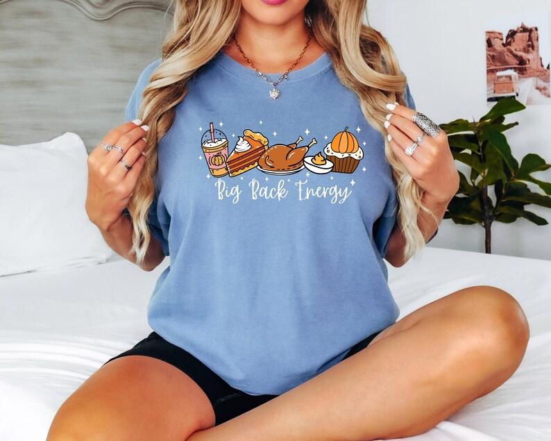 Puede incluir: Camiseta azul claro con un gr&aacute;fico de alimentos de Acci&oacute;n de Gracias, que incluye un caf&eacute; con leche de calabaza especiada, tarta, pavo y una magdalena. El texto "Big Pack Energy" est&aacute; debajo de las ilustraciones de comida. La camiseta es de material suave.