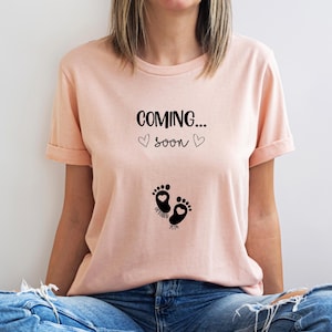 Op de afbeelding: Een perzikkleurig t-shirt met korte mouwen en zwarte tekst COMING... SOON met twee kleine hartjes. Onder de tekst staan twee zwarte babyvoetjes, elk met een klein hartje erin, en de woorden OCTOBER en 2019.