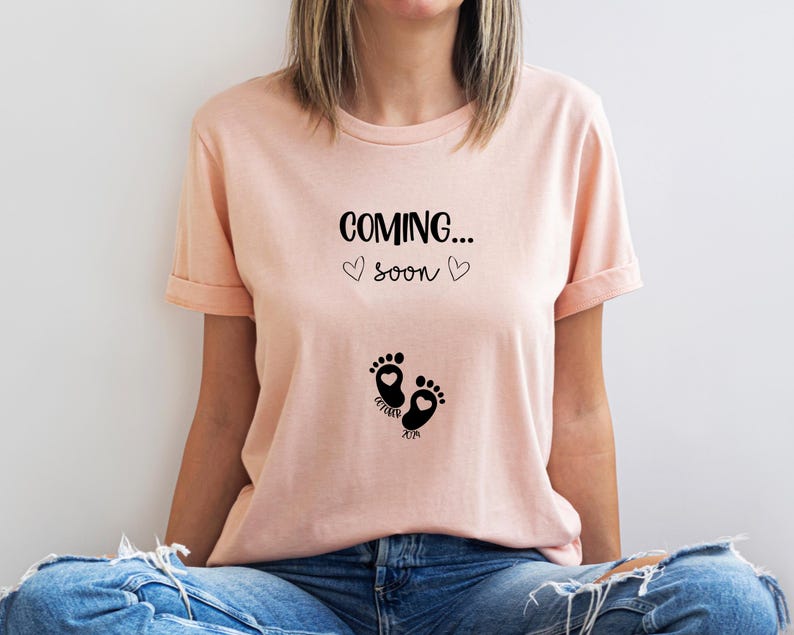 Op de afbeelding: Een perzikkleurig t-shirt met korte mouwen en zwarte tekst COMING... SOON met twee kleine hartjes. Onder de tekst staan twee zwarte babyvoetjes, elk met een klein hartje erin, en de woorden OCTOBER en 2019.