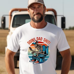 May include: White t-shirt with a graphic of a skeleton wearing a baseball cap and sitting on a toilet, using a phone. The text "DOING DAD SHIT!" is above the graphic. The background includes a truck and a field.