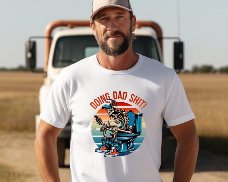 May include: White t-shirt with a graphic of a skeleton wearing a baseball cap and sitting on a toilet, using a phone. The text "DOING DAD SHIT!" is above the graphic. The background includes a truck and a field.